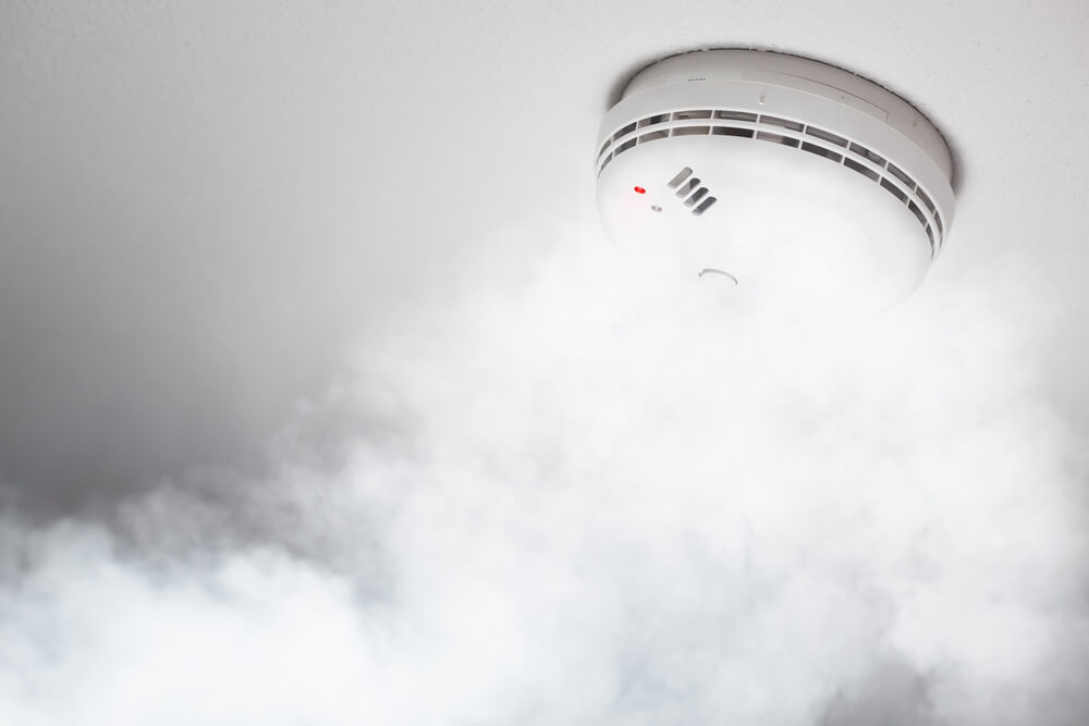 Smoke billowing around a smoke detector on a ceiling.