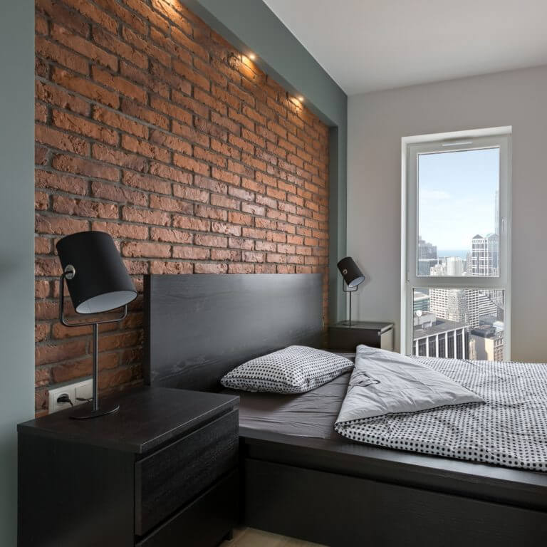 Een bakstenen muur achter een bed in een slaapkamer in industriële stijl.