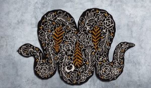 An image shows an artistic design for a rug in the shape of a snake, with white and golden brown embroidery on a black surface.