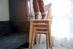 A person seen from the legs down is standing on their tip-toes on top of a stack of chairs, trying to reach something.