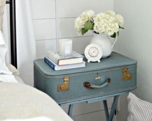 A picture showing a vintage suitcase being used as a nightstand beside a bed, with a couple books and a vase on top of it.