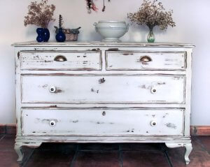 A vintage dresser with flaking white paint is in the hallway to a house, with various flower vases on top of it.
