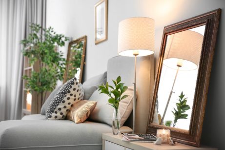 A room with light colored furniture, a lamp, mirrors, and plants.