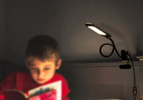 A boy reading a book under the light of his flexible lamp.