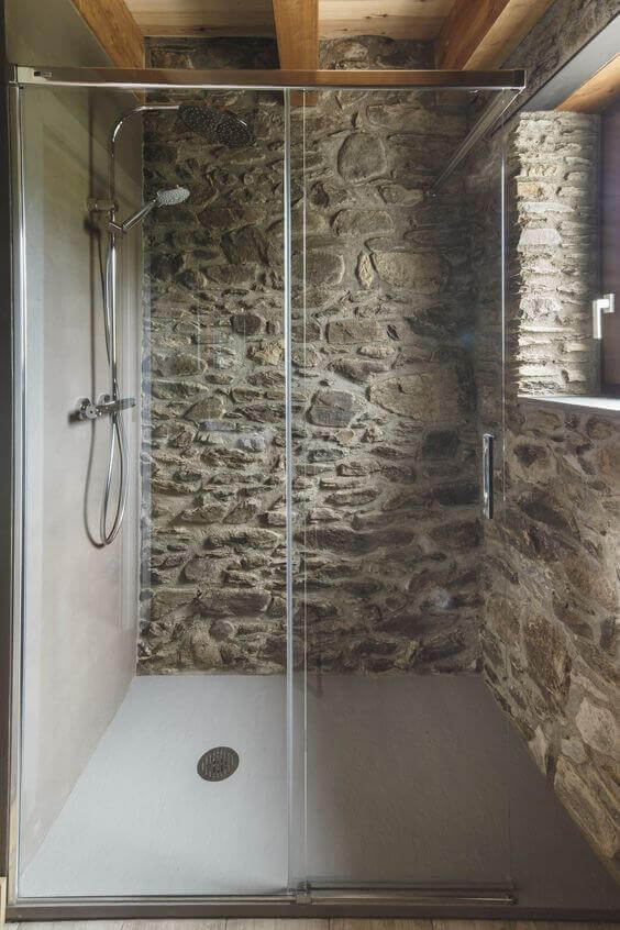 A shower with a glass door in a stone bathroom with a wooden ceiling.